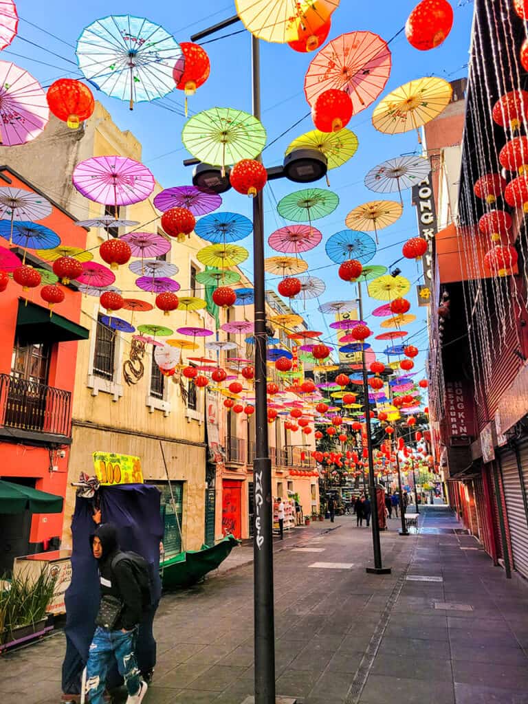 A man pulls a street stall through Barrio Chino in Mexico City with paper umbrellas and lanterns hanging overhead.