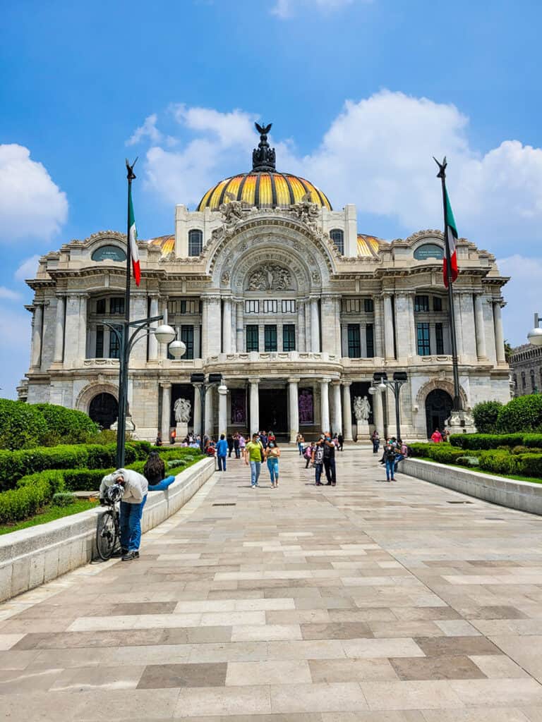 The golden dome of the Palacio de Bellas Artes glistens in the sun under a blue sky with fluffy white clouds.