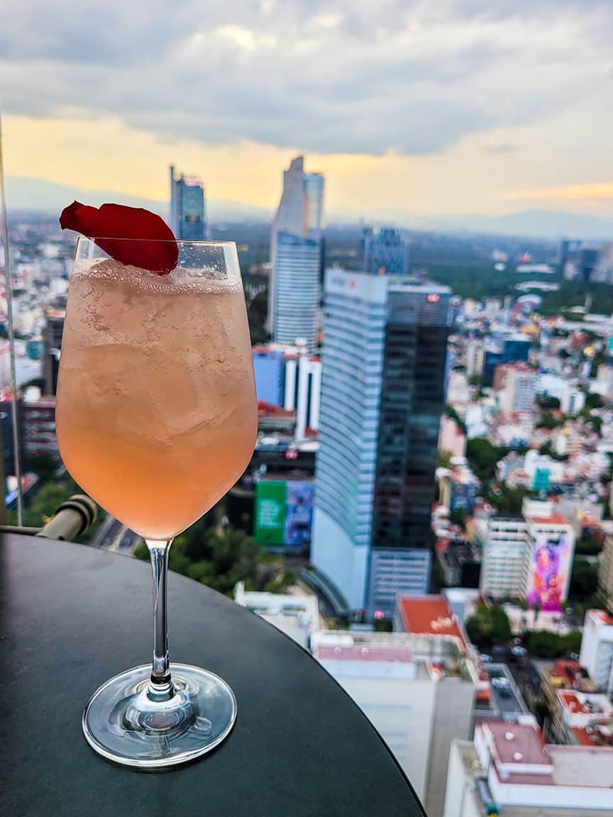 The Montparnasse cocktail topped with a single red rose petal on a table at Cityzen Rooftop Kitchen with a Mexico City view in the background.