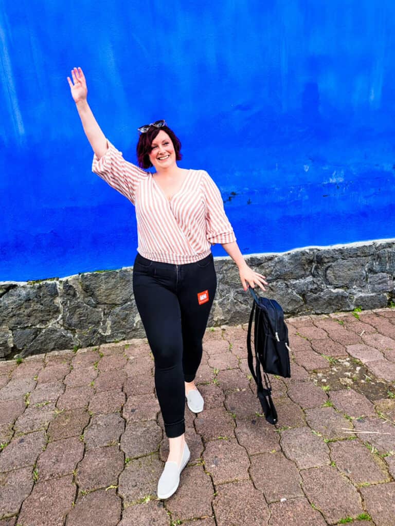 Ashlea smiles as she stands in front of the blue walls of Frida Kahlo House in Mexico City with one arm raised.