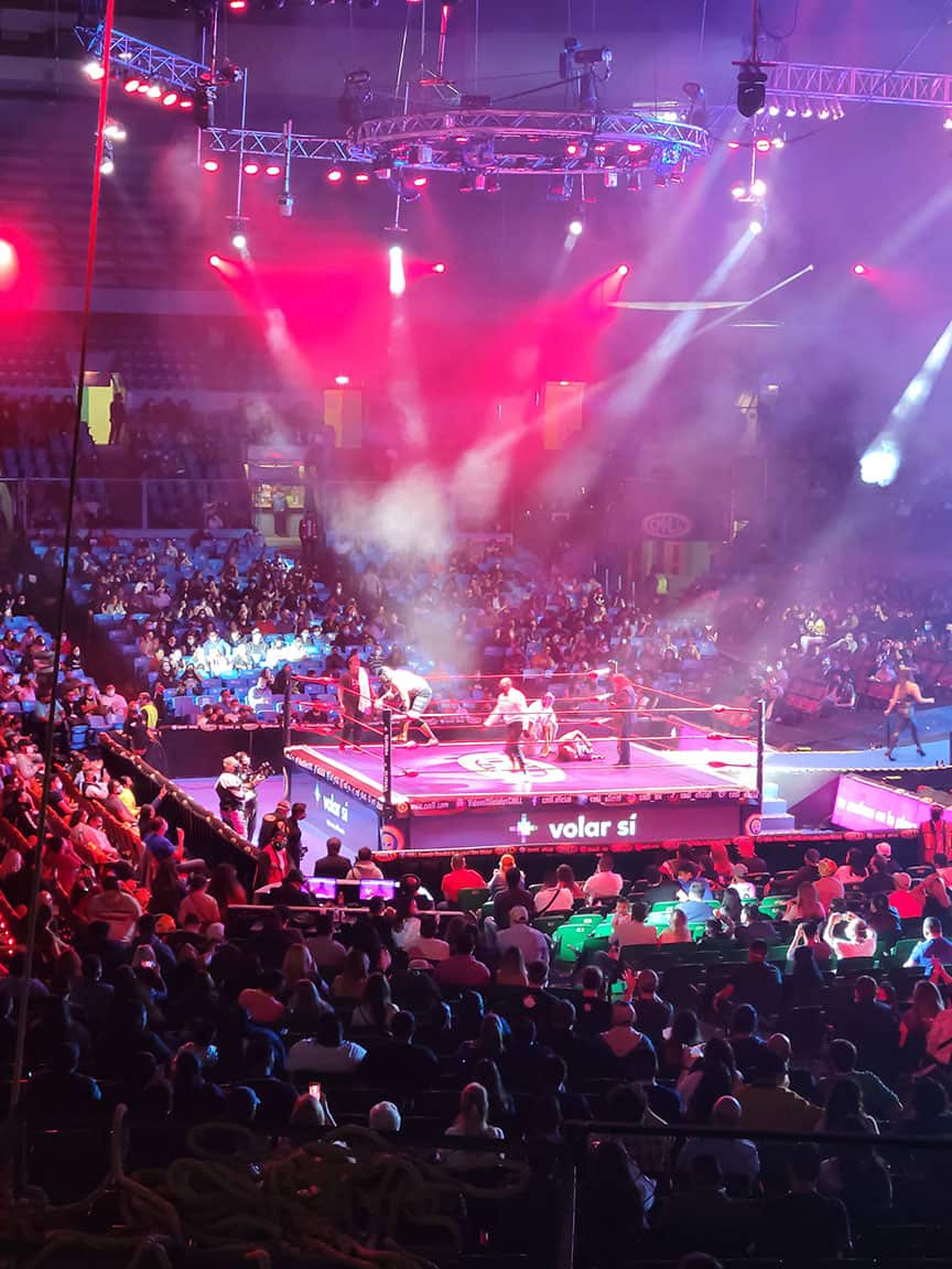 A crowd watches a Lucha Libre match under bright lights while the wrestlers prepare for their next move.
