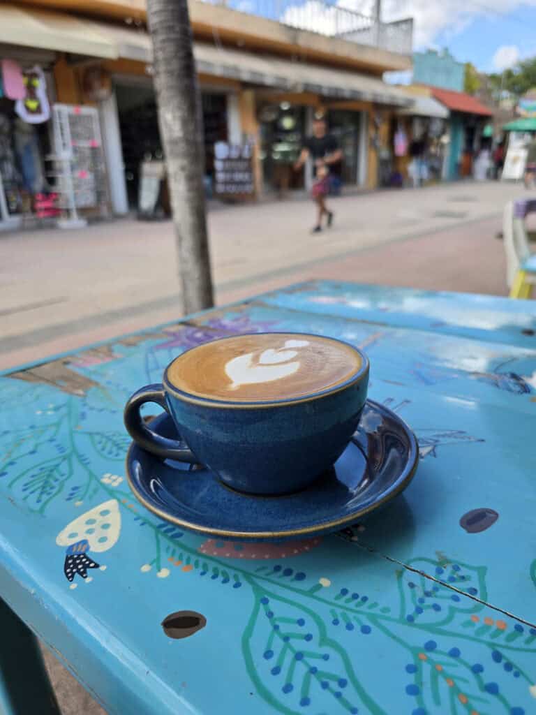 A flat white coffee at Coz Coffee Roasters in Cozumel with shops and a man walking by in the background.